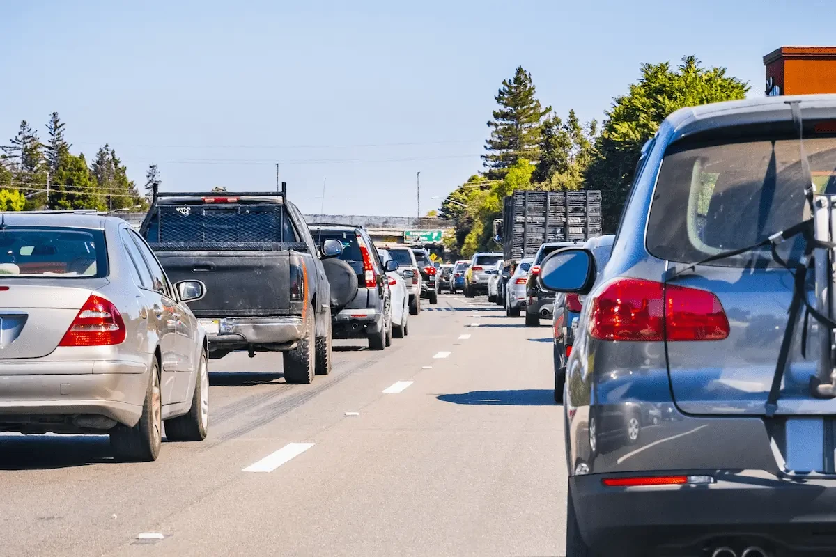 Rear view of traffic on a multilane freeway on a sunny day, with a silver sedan at the left edge, a dark pickup truck with a mesh rear window guard, a gray SUV with a rear-mounted rack at the right, and a line of cars and a truck carrying stacked cages ahead, with trees, an overpass, and a green highway sign in the distance.