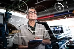 Collision repair technician using a tablet in a shop with digital interface overlays representing AI tools and data-driven workflows.
