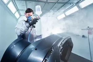 A technician in a paint booth sprays clear coat onto a large vehicle body panel while wearing protective gloves, coveralls, and a respirator, with paint mist visible in the air.