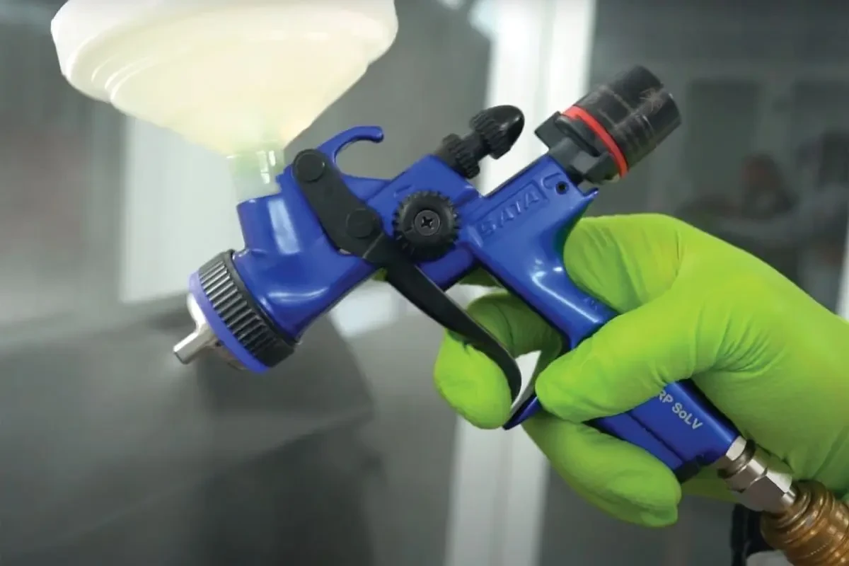 Close-up of a painter's gloved hand holding a blue SATA spray gun with an attached gravity-feed cup while spraying a panel in a paint booth.