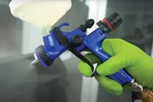 Close-up of a painter's gloved hand holding a blue SATA spray gun with an attached gravity-feed cup while spraying a panel in a paint booth.