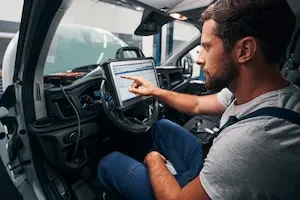 A mechanic in coveralls sits in a vehicle's driver seat, pointing at a mounted diagnostic screen displaying data readouts, with cables connected to the dashboard.