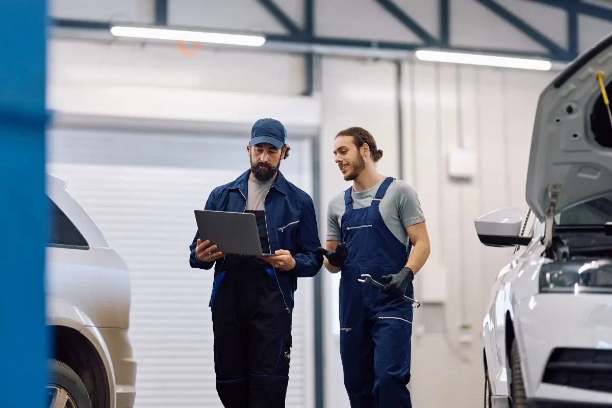 Two automotive technicians reviewing information on a laptop in a collision repair shop bay with vehicles on either side.