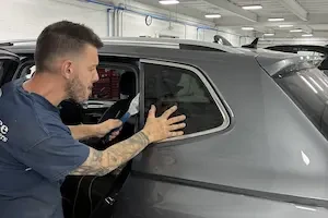 Man with beard and tattoos prying auto glass out of the side of a gray vehicle