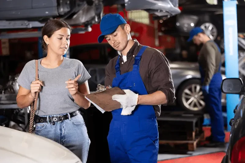 A woman in a gray T-shirt and jeans speaks with a mechanic in a blue cap and blue overalls holding a clipboard inside an auto repair shop, with vehicles on lifts and another mechanic working in the background.