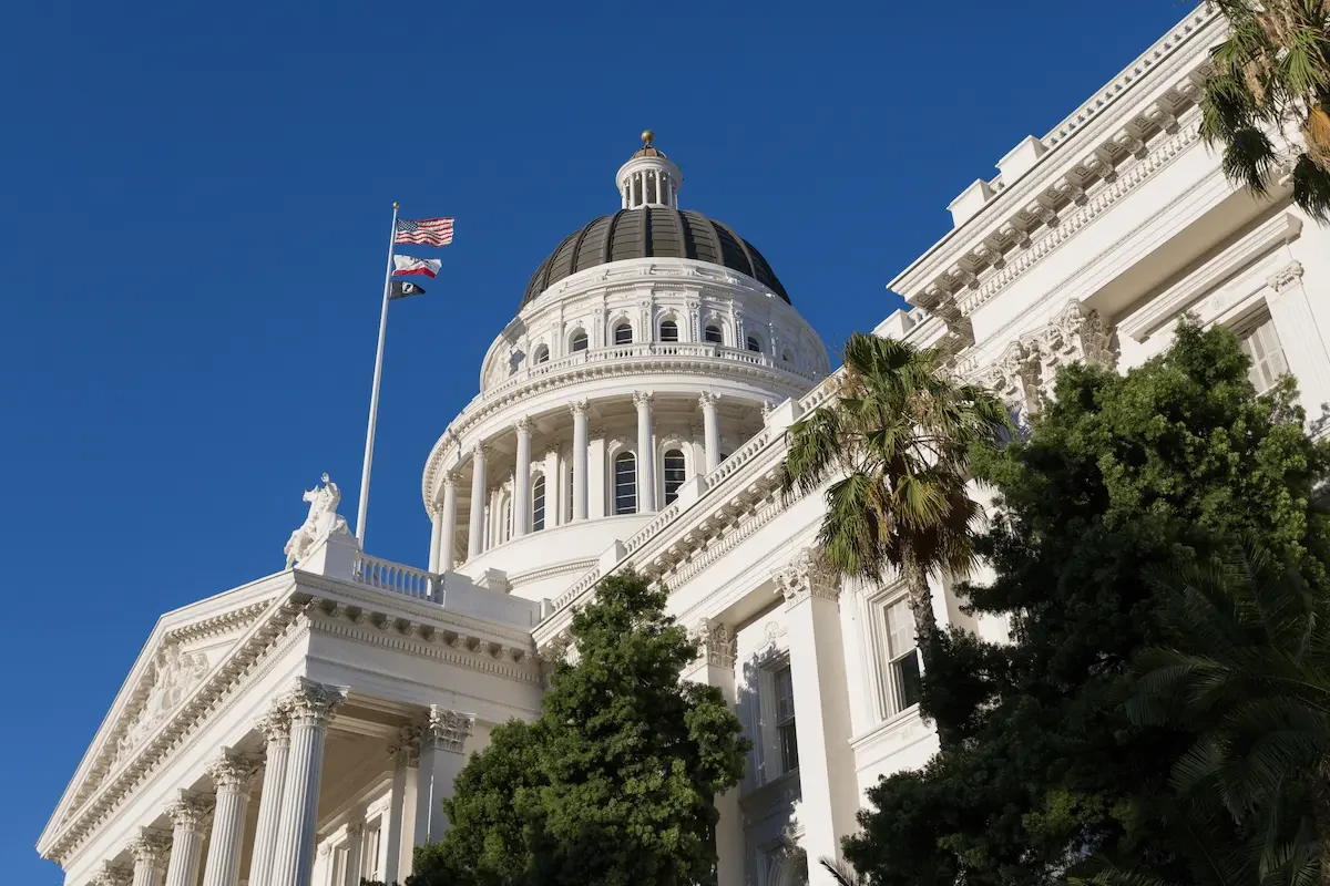 Low-angle view of the California State Capitol dome and facade, with white Corinthian columns, an American flag and California state flag on a pole in the foreground, palm trees and other trees along the base of the building, against a clear blue sky.