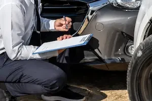A person in business attire kneeling beside a damaged silver car, holding a clipboard marked insurance and pointing a pen at the vehicle's front bumper.