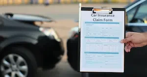 A hand holds a clipboard with a car insurance claim form, with two vehicles parked in the background.
