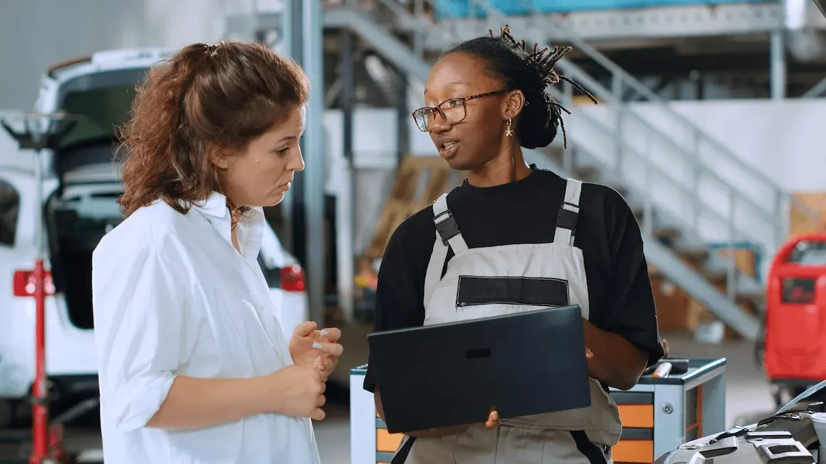 A woman in overalls and glasses holds a laptop while speaking with a woman in a white shirt in an auto repair shop.