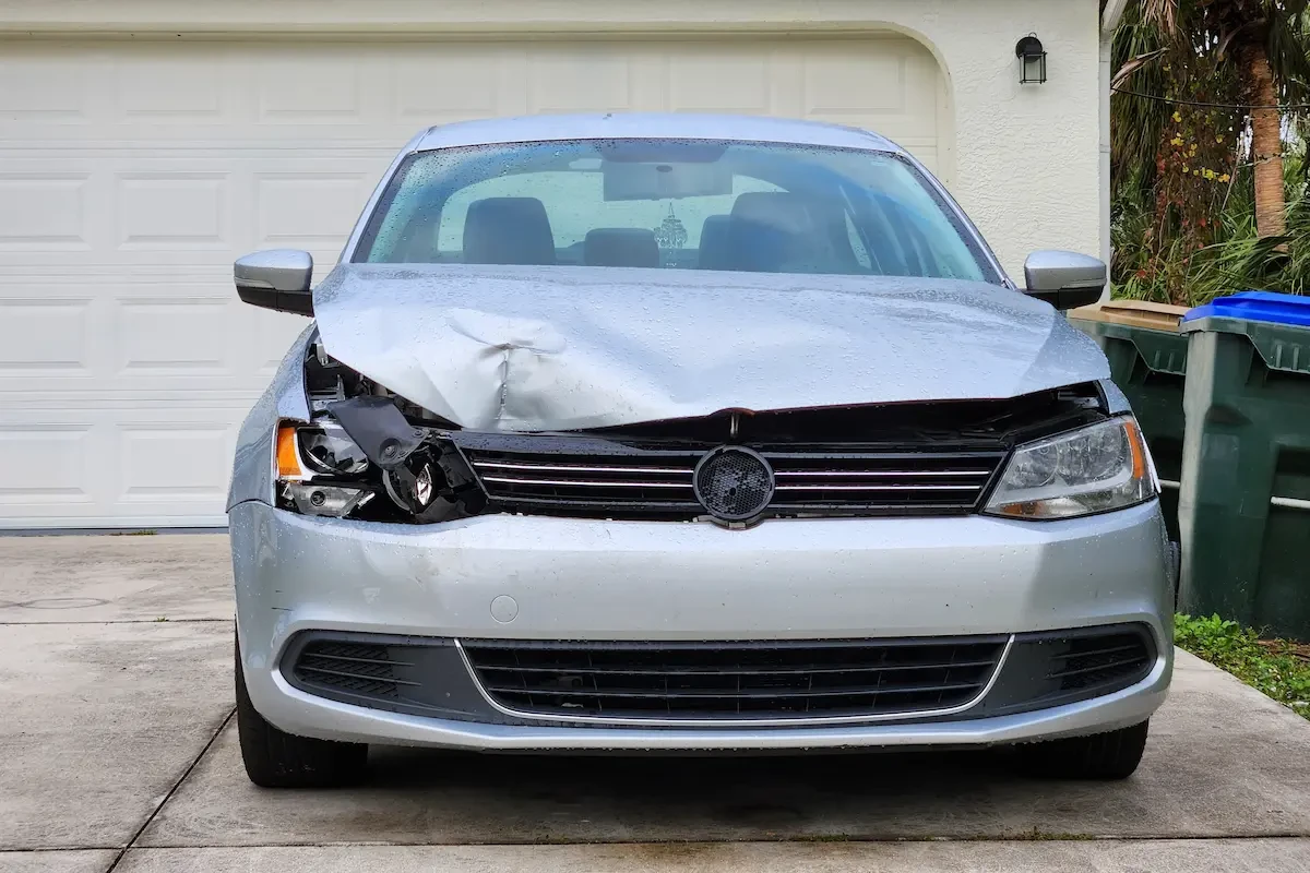 A silver sedan with a dented hood and damaged front bumper and headlight parked in a driveway in front of a closed garage door, with trash bins and palm trees visible in the background.