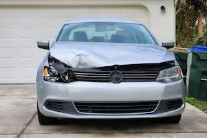 A silver sedan with a dented hood and damaged front bumper and headlight parked in a driveway in front of a closed garage door, with trash bins and palm trees visible in the background.