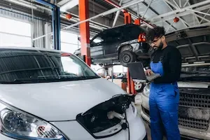 Mechanic in blue coveralls running a diagnostic scan from a laptop connected to a vehicle's engine bay, with additional cars on lifts in the background.