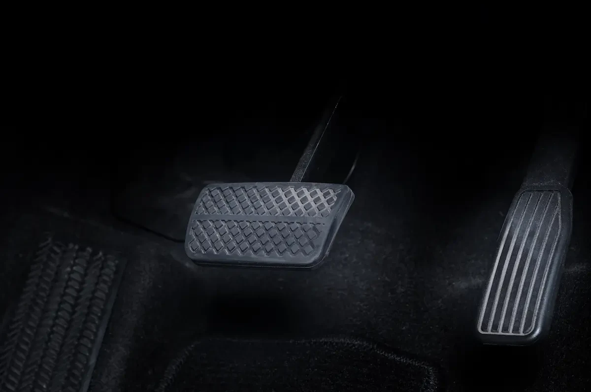 Close-up of a car's brake and accelerator pedals on a dark carpeted floor mat, viewed from an angle showing the pedals' textured rubber surfaces.