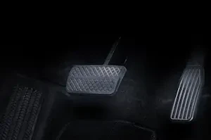 Close-up of a car's brake and accelerator pedals on a dark carpeted floor mat, viewed from an angle showing the pedals' textured rubber surfaces.