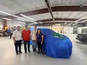 Four people standing in a shop in front of a car covered in a tarp with a bow on it