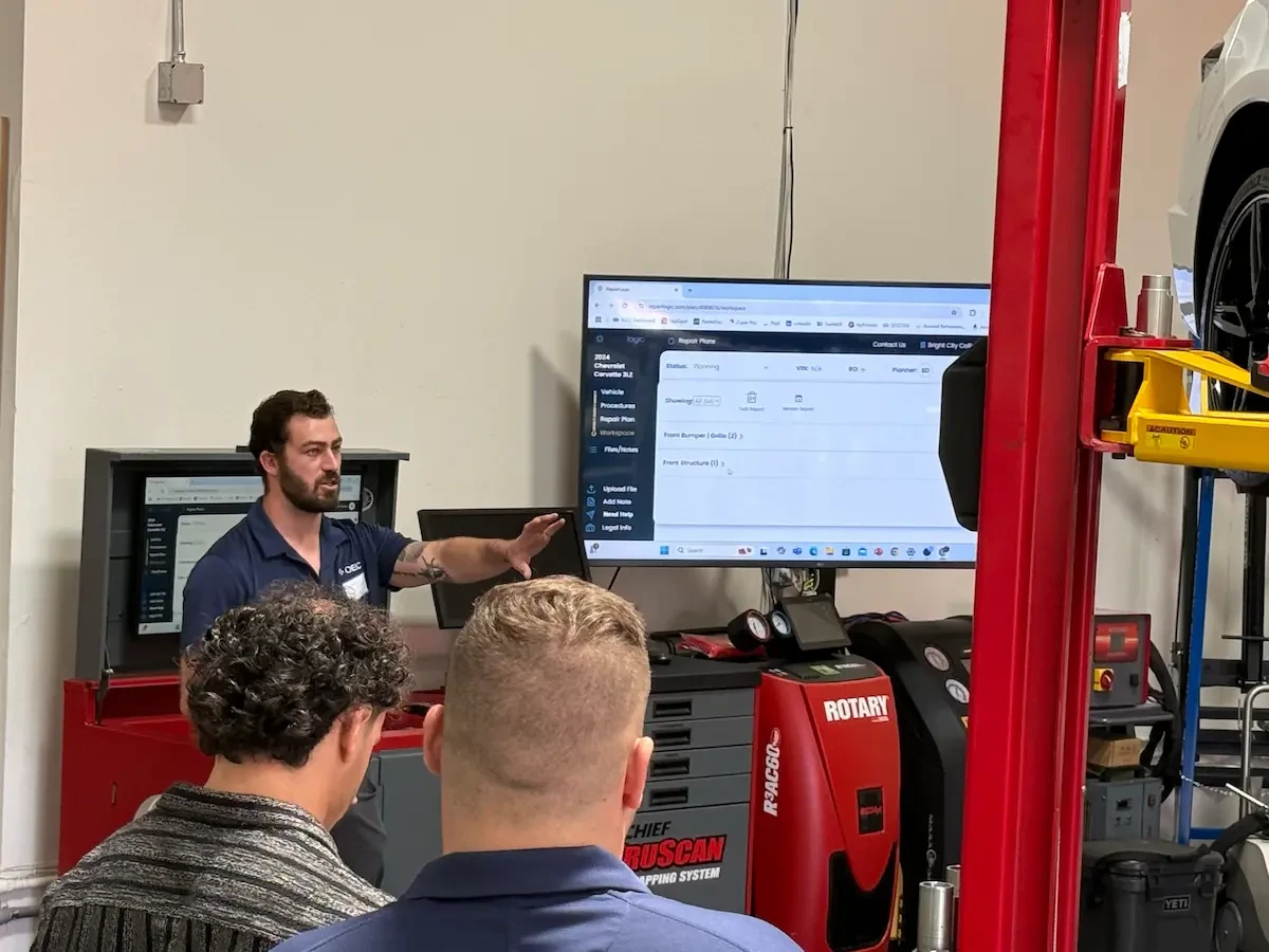 A man in a navy OEC polo shirt gestures toward a large wall-mounted monitor displaying the RepairLogic software interface, showing a repair plan for a 2024 Chevrolet Corvette with sections for Front Bumper/Grille and Front Structure. Two other men, seen from behind, watch the screen. The shop setting includes a red two-post lift, a Rotary R3ACG0 AC recharge machine, a Chief Truscan mapping system cart, and various shop equipment and tool storage.