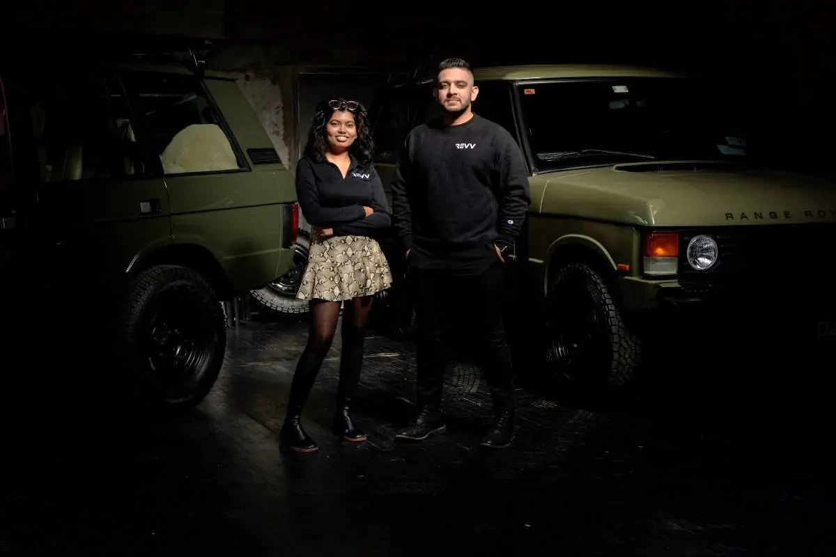 Two people in black Revv-branded sweatshirts stand in front of a green Range Rover in a dark garage.