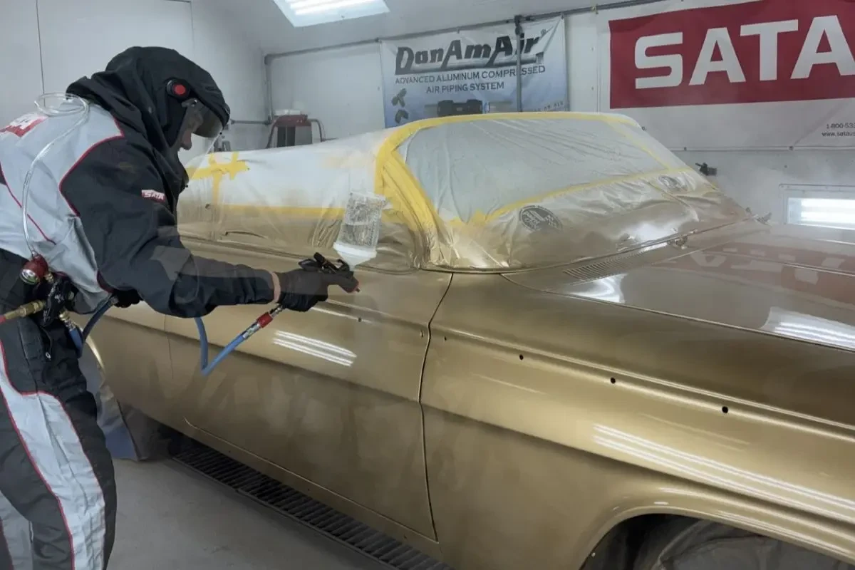A painter wearing a SATA supplied-air respirator hood and protective suit sprays gold basecoat onto a masked classic car in a spray booth, with SATA and DanAmAir banners on the wall behind.