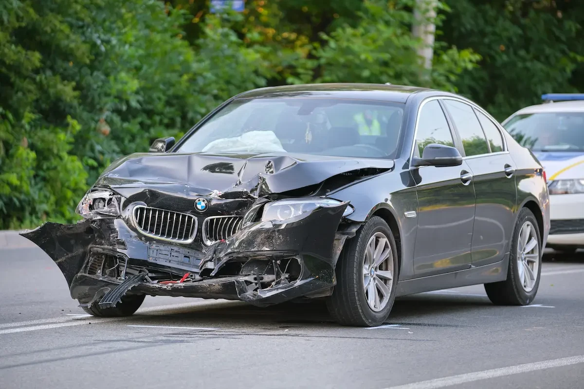 A dark gray BMW sedan with a crushed, torn-open front end and a deployed airbag sits on a roadway, with a police vehicle parked behind it.