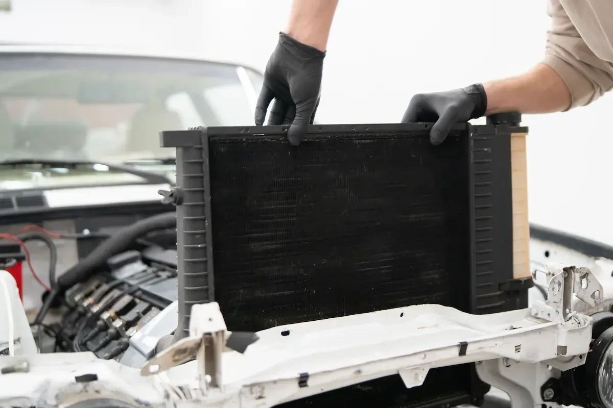 A mechanic wearing black gloves lifts a radiator out of a car's engine bay during a repair.