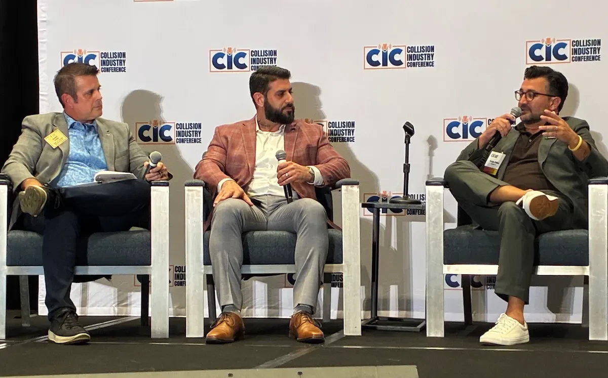 hree men sit in chairs on a stage with microphones in front of a repeating Collision Industry Conference backdrop; the man on the left holds a notepad, the man in the center wears a plaid blazer and speaks into a microphone, and the man on the right wears a green suit and gestures while speaking.