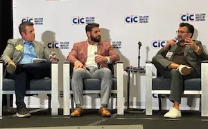 hree men sit in chairs on a stage with microphones in front of a repeating Collision Industry Conference backdrop; the man on the left holds a notepad, the man in the center wears a plaid blazer and speaks into a microphone, and the man on the right wears a green suit and gestures while speaking.