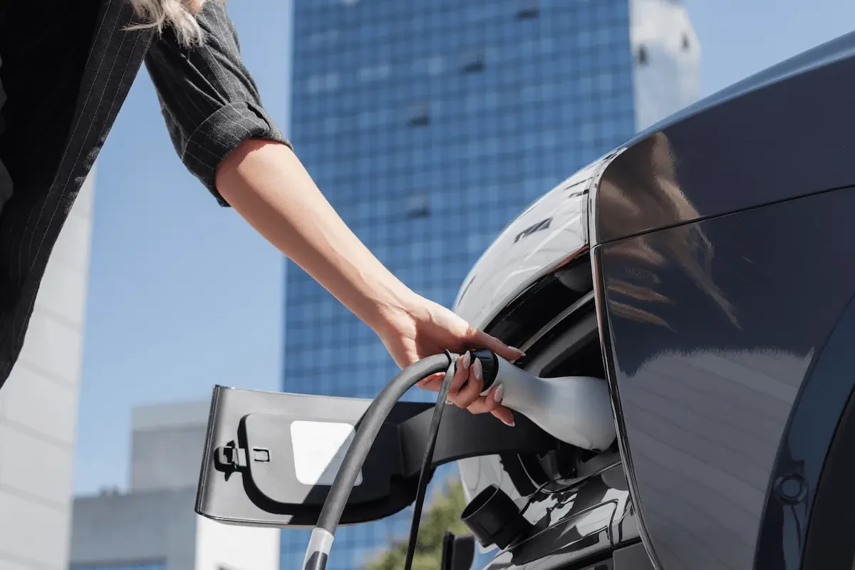Woman's hand holding a charging plug inserted into the charging port of a dark gray car, with a blue glass office building in the background.