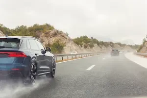A rear view of several vehicles, including a dark SUV in the foreground, driving on a wet mountain highway with tire spray visible.