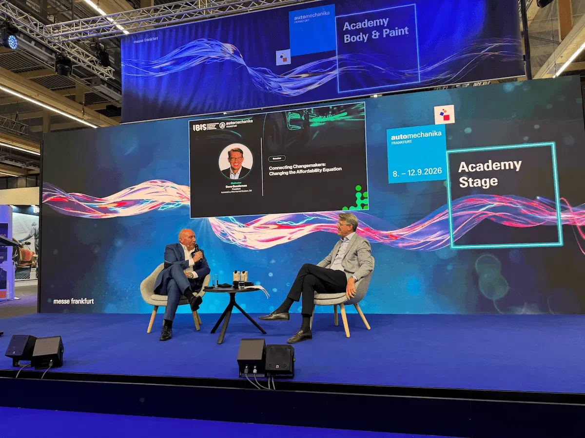 Two men in suits sit in gray armchairs on a blue-carpeted stage, facing each other across a small black round table holding water glasses and bottles. The man on the left speaks into a handheld microphone. Behind them, a large screen displays a headshot and the text "Connecting Changemakers: Changing the Affordability Equation" beside IBIS and Automechanika Frankfurt logos. Signs to the right read "Academy Stage" and "8. – 12.9.2026," and a banner above reads "Academy Body & Paint."