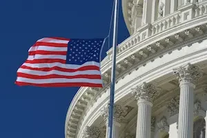 American flag on a pole in front of the U.S. Capitol dome, with white Corinthian columns visible against a blue sky.