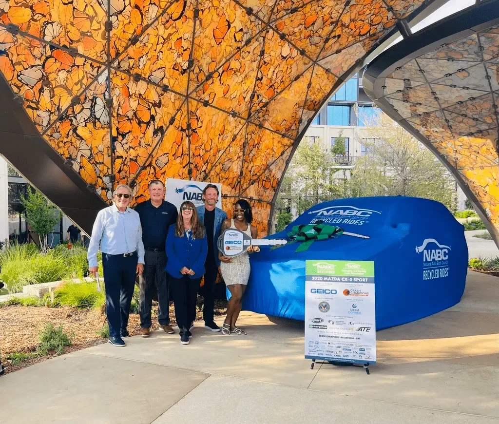 Five people stand beneath a canopy decorated with orange butterfly artwork. One woman holds an oversized GEICO key. Beside them is a vehicle under a blue NABC Recycled Rides cover with a green bow, next to a sign reading "2020 Mazda CX-5 Sport" with sponsor logos including GEICO, Crash Champions, and CIECA CONNEX.