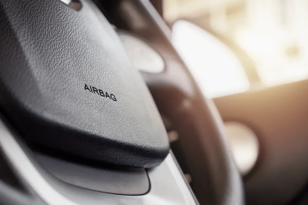 A close-up of a black textured steering wheel airbag cover embossed with the word "AIRBAG," with the steering wheel rim and a blurred, sunlit vehicle interior in the background.