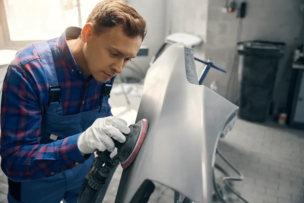 A technician in plaid shirt, blue overalls, and white gloves uses an orbital sander on a gray vehicle panel mounted on a stand in a repair shop.