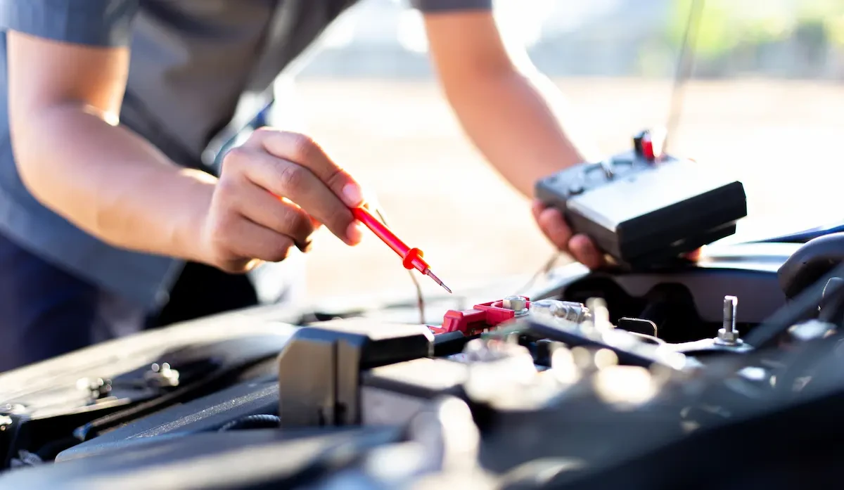 Close-up of a technician's hands holding a multimeter and touching a red test probe to a vehicle battery terminal under the hood.