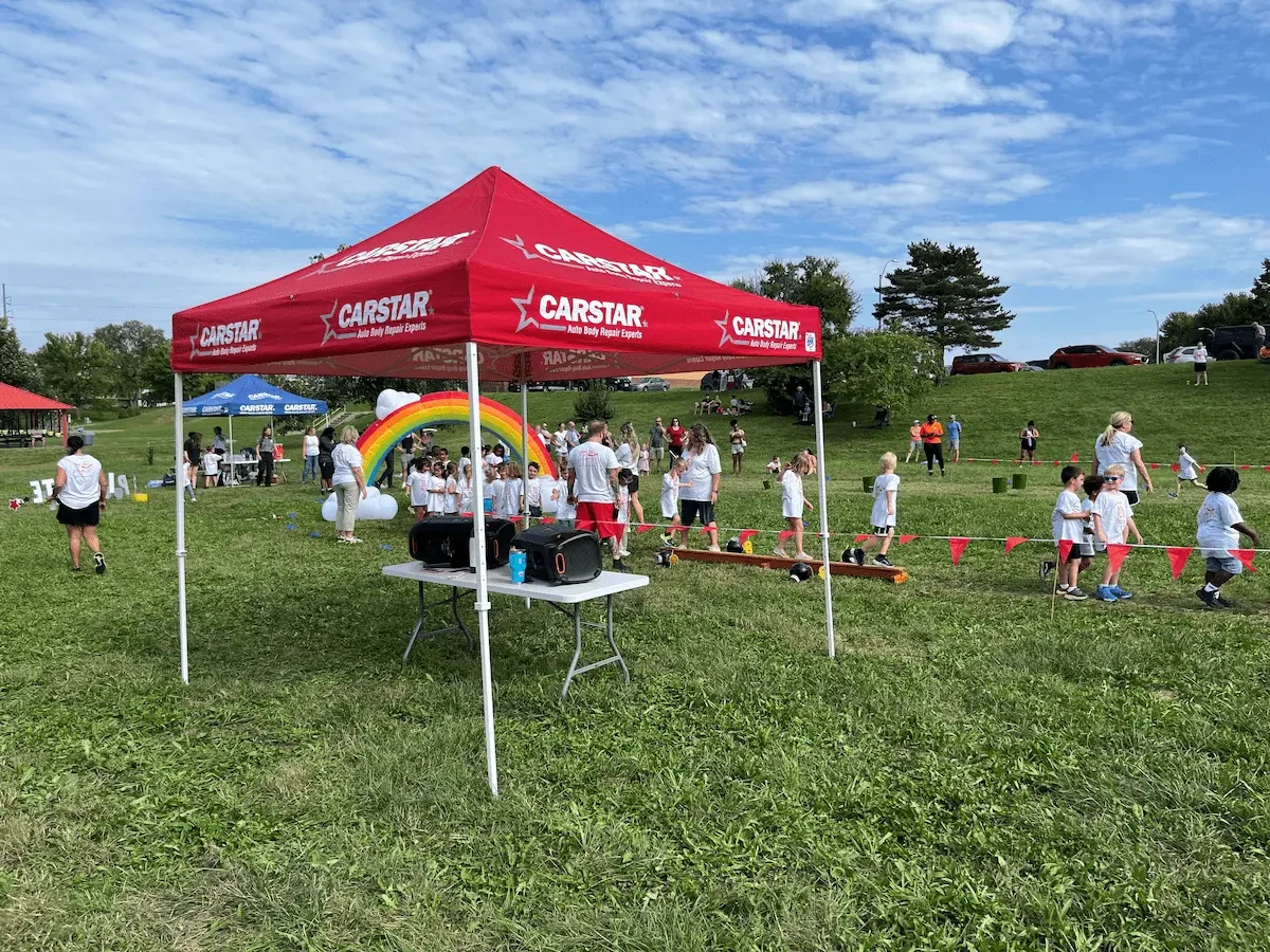 A red CARSTAR canopy tent stands on a grassy field under a blue sky with scattered clouds. Under the tent is a table with supplies and a rainbow balloon arch. Children and adults in white shirts stand near the tent and along a row of small orange cones, with trees and a picnic shelter in the background.