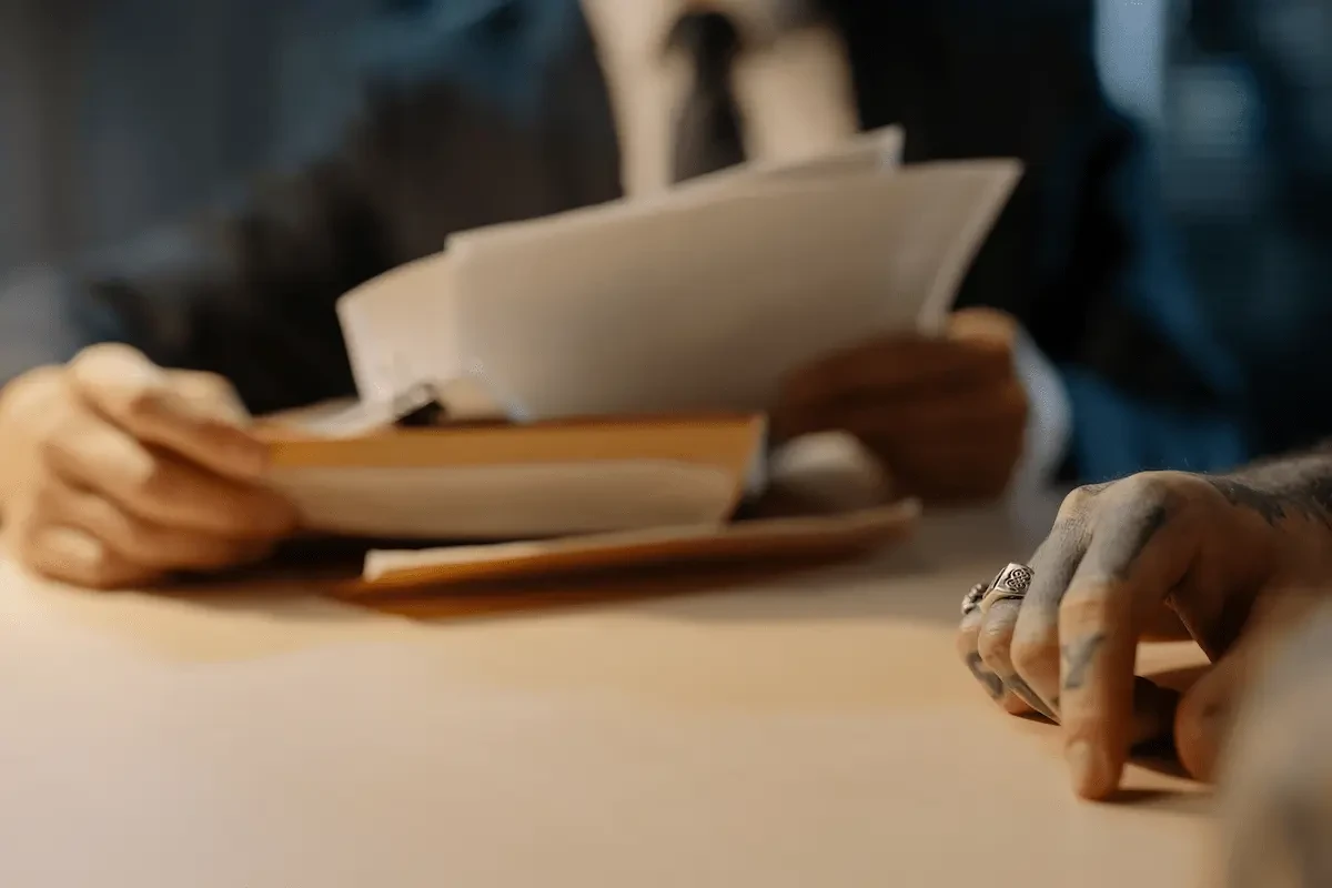 A blurred person in a dark suit, white shirt, and dark tie holds a stack of white papers and a tan folder with a black binder clip above a light wood table. In the right foreground, a tattooed hand wearing silver rings rests on the table's edge, in sharp focus.
