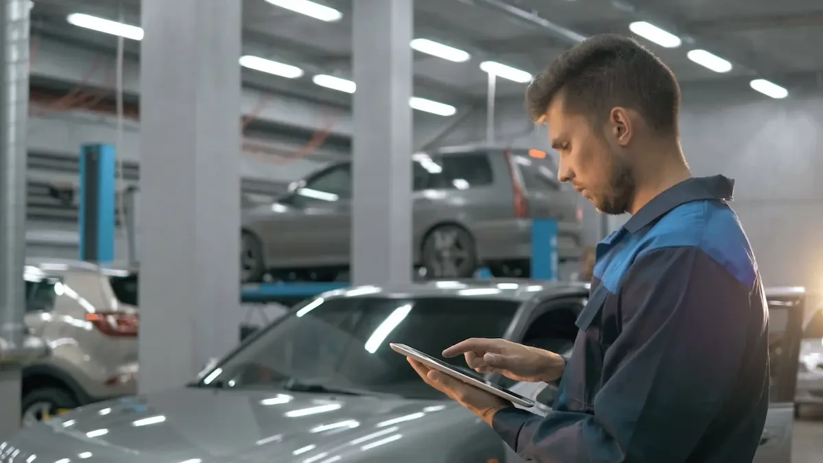 Man in navy coveralls with light blue shoulder panels stands in profile in an auto repair garage, using his index finger on a tablet he holds in both hands, with cars on lifts and industrial lighting in the background.