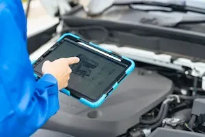 A mechanic in a blue coverall points at a technical diagram on a rugged tablet held in front of an open vehicle engine bay.