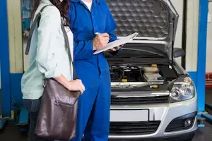 A mechanic in blue coveralls writes on a clipboard beside a customer holding a shoulder bag, standing next to a silver sedan with its hood raised in a repair shop.
