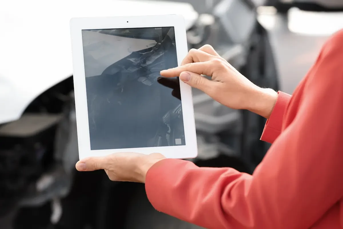A person in a coral top holds a white tablet, photographing the open engine bay or undercarriage of a vehicle.
