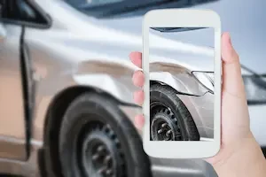A hand holds a smartphone up to a damaged silver car, with the phone screen showing a close-up photo of a dented front fender and wheel.