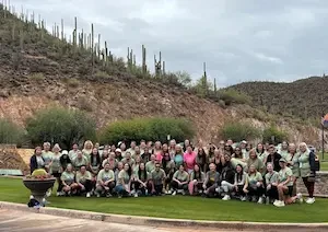 Large group of women in matching light green T-shirts posed on a lawn for a group photo, with a desert hillside covered in saguaro cacti in the background and a stone resort sign partially visible at left.