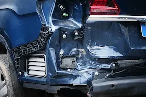 The rear bumper and quarter panel of a dark blue car are crumpled and torn open from a collision, exposing wiring and structural components underneath.