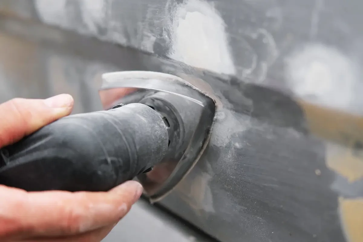 Close-up of a gloved hand holding a rotary grinder against a metal seam on a vehicle body panel, with dust and debris visible.