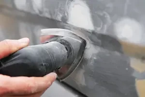Close-up of a gloved hand holding a rotary grinder against a metal seam on a vehicle body panel, with dust and debris visible.