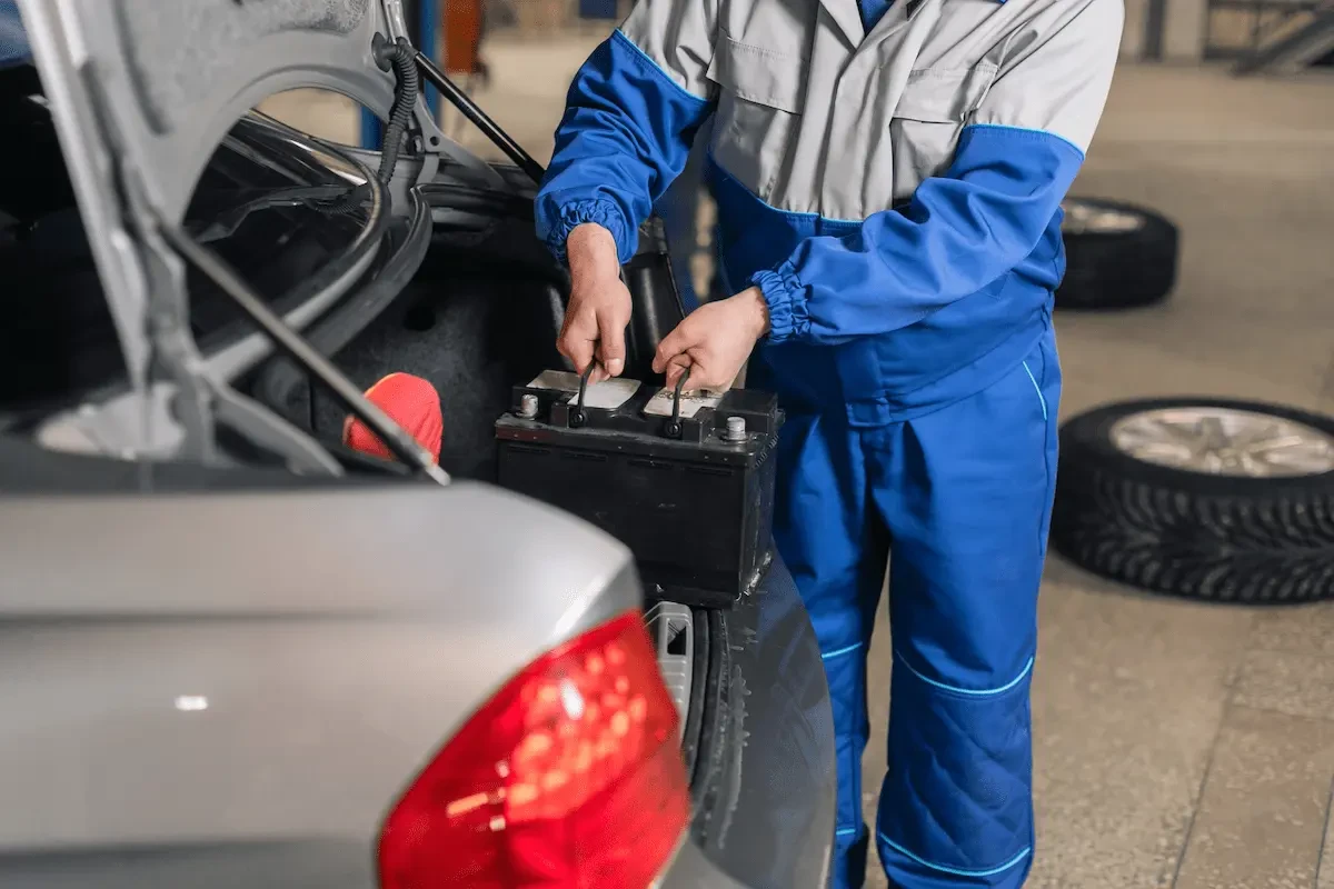 A mechanic in blue and grey coveralls disconnects a car battery mounted in the trunk of a silver sedan, with stacked tires visible in the shop background.
