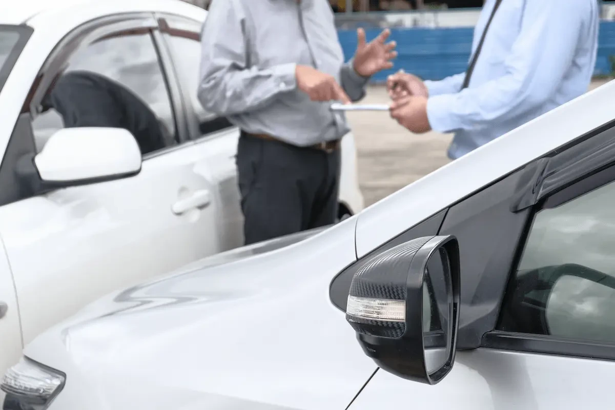 Two people in business attire stand between two white cars in a parking lot, one holding a notepad and pen while gesturing during a conversation; the car on the left has a visible dent near the front door.