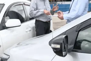 Two people in business attire stand between two white cars in a parking lot, one holding a notepad and pen while gesturing during a conversation; the car on the left has a visible dent near the front door.