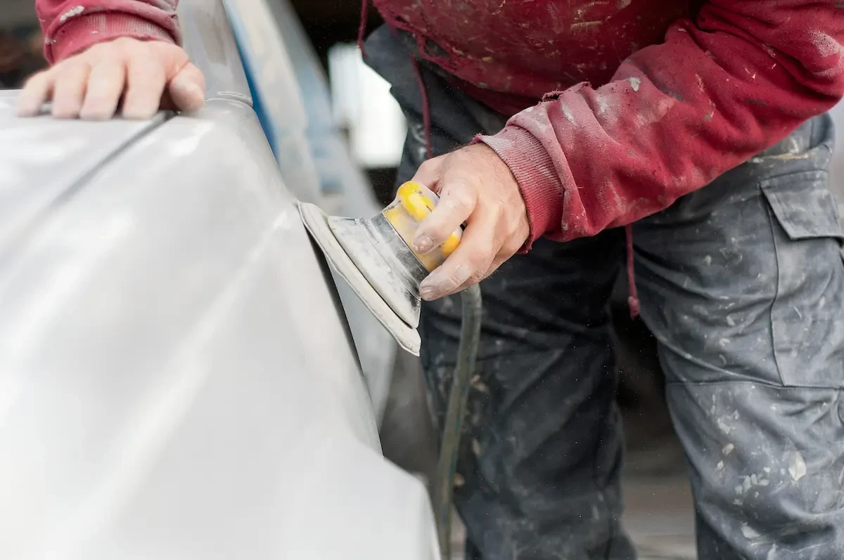 A worker's gloved hand uses a yellow hand sander on the primer-gray body panel of a vehicle, wearing a maroon long-sleeve shirt and gray work pants marked with dust and residue.