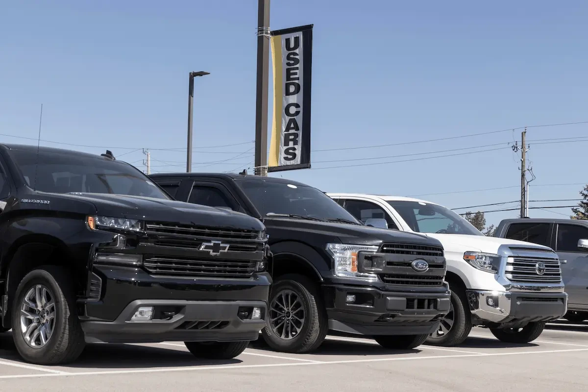 A row of used cars in a lot under a used cars sign. There are two large black SUVs or trucks, a white one, and a silver one.